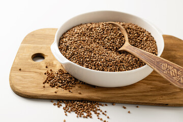 Dry uncooked buckwheat groats in a white dish, wooden board, spoon, on a white background, close-up