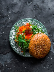 Breakfast burger with egg, salmon, arugula and red caviar on a dark background, top view