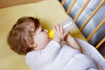 Cute little baby girl holding bottle with formula mild and drinking. Child in baby cot bed before sleeping