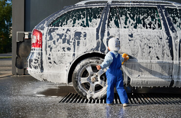 Rear view of toddler boy in jumpsuit and cap, standing with sponge in his hands near side door of car body covered with soap suds. Child rubbing soap suds, taking part in cleaning auto at the car wash