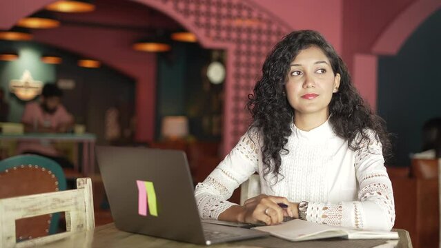positive Portrait of young Indian girl siting at table in the restaurant. effectual Asian business woman smiling working and typing on laptop computer and writing a notebook. looking at laptop screen.