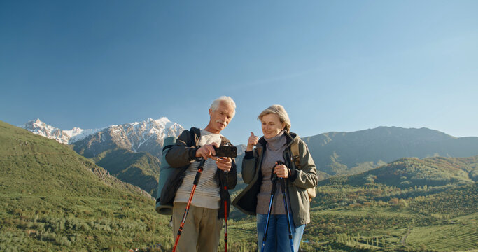 Mature caucasian couple travelling together, having a nordic walking hike in spring mountains, then stopping to take a picture on smartphone, spending time after retirement - recreational pursuit