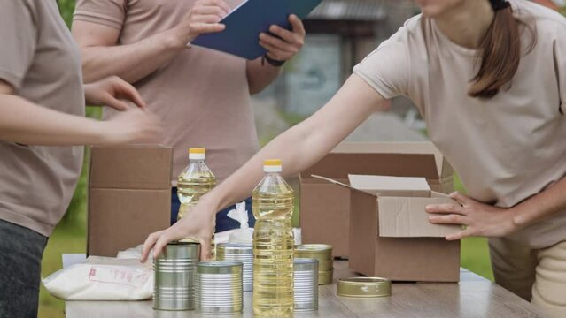 Close-up of unrecognizable members of charity organization pack canned and non-perishable products in cardboard boxes outdoors. Charity, donation and volunteering concept, global food crisis