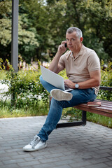 Vertical confident, successful mature gray haired entrepreneur use laptop and mobile phone on bench and look at camera