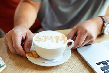 Cropped hands hold coffee cup with cream foam SMM of cinnamon with cookies on wooden cafe table. Social Media Marketing
