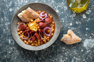 Plate of chorizo and chickpea stew served with crusty bread on a dark-brown granite background, horizontal shot, top view