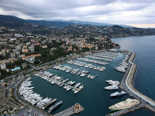 Fototapeta premium Aerial view of Sanremo, Italian city on the seashore in Liguria, north Italy. Drone flying along the port over beaches and boardwalk with palm trees and Birds Eye of yacht parking in San Remo, Italy.