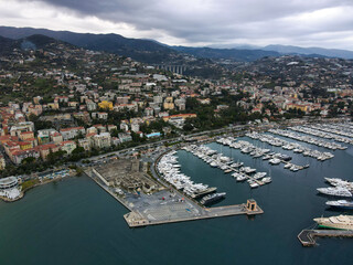 Fototapeta premium Aerial view of Sanremo, Italian city on the seashore in Liguria, north Italy. Drone flying along the port over beaches and boardwalk with palm trees and Birds Eye of yacht parking in San Remo, Italy.