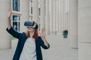 Young ginger woman dressed formally stands outdoors and using VR headset