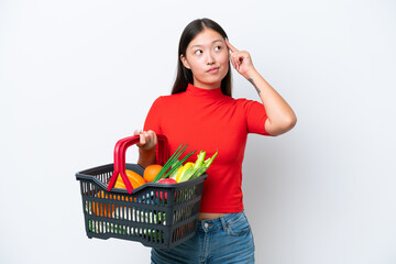 Plakat Young Asian woman holding a shopping basket full of food isolated on white background having doubts and thinking