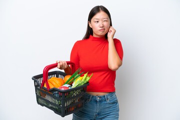 Obraz na płótnie Canvas Young Asian woman holding a shopping basket full of food isolated on white background frustrated and covering ears