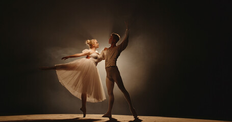 Professional duet of ballet dancers performing on stage isolated on smoked black background. Young...