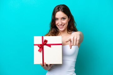 Young caucasian woman holding a gift isolated on blue background points finger at you with a confident expression