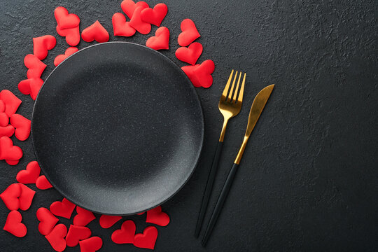 Valentines day romantic table setting. Empty or closeup of a dinner black plate, knife, fork and decorative silk hearts on black background. Holiday concept. Copy space for inscriptions.