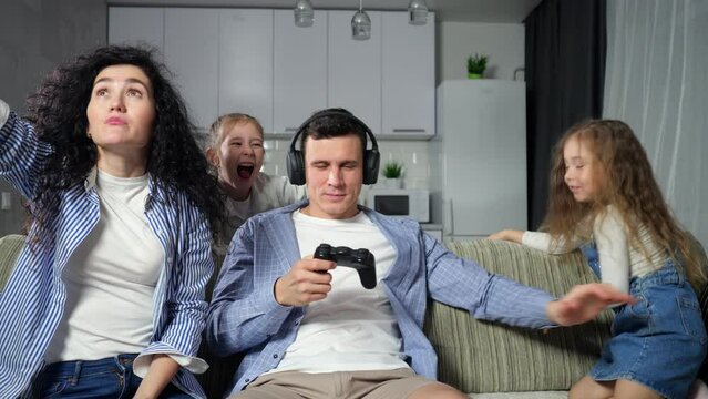 Insistent daughters and wife try to get attention of father playing games with controller. Man enjoys playing games and puts on headphones to distract