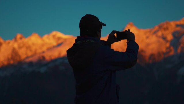 Silhouette shot of Indian male hiker taking photo of snow covered Himalayan mountains during sunset for his social media during his vacation in winter season at Manali in Himachal Pradesh, India. 
