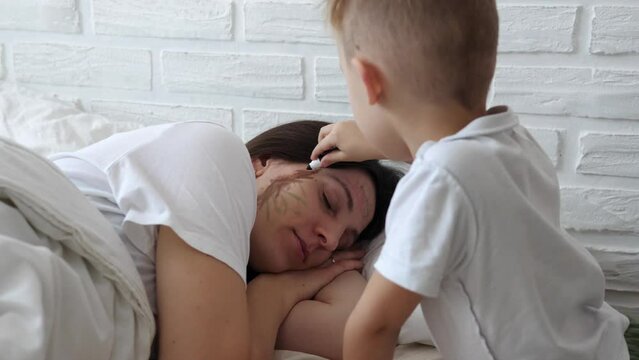Little naughty charming boy draws markers on the face of a sleeping mom.