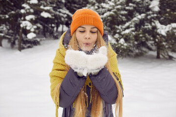 Fototapeta premium Winter mood. Portrait of beautiful young woman blowing snow from her palms during winter walk. Snowflakes fly from hands of girl in winter wear who walks in snowy forest and enjoys winter frosty day.