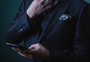 Smart young man in luxury black suit using smartphone on dark green background.