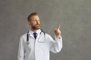 Serious man doctor showing aside pointing with finger at grey empty copy space. Confident male medical worker in white coat and stethoscope thinking, brainstorming about question