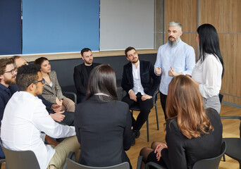 Business team sitting in circle and discussing. Professional male leader and coach who training staff speaks at meeting with various managers. Serious business people sitting on chairs in circle.