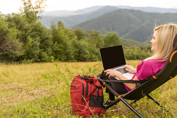 A woman in a sports warm suit works on a laptop outdoors in a mountainous area.