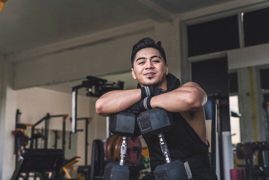 A confident asian man smirking while leaning on dumbbells placed on his knees at the gym. Wearing a black hooded low cut tank top and gloves. Weight training at the gym.