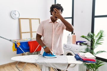 African man with curly hair ironing clothes at home peeking in shock covering face and eyes with...