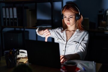 Young caucasian woman working at the office at night looking unhappy and angry showing rejection and negative with thumbs down gesture. bad expression.