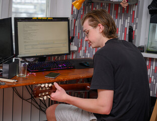 A young guy learns to play the guitar sitting at a table at home online on a computer.