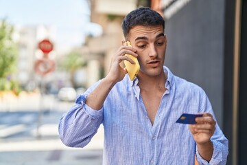 Young hispanic man talking on the smartphone and using credit card at street