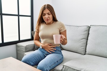 Young hispanic woman with stomatchache drinking tea at home