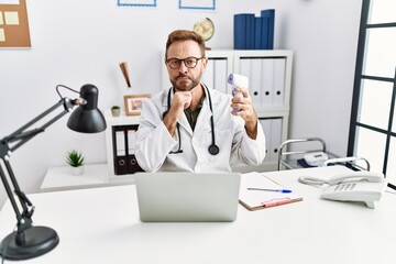 Obraz premium Middle age doctor man holding thermometer at the clinic serious face thinking about question with hand on chin, thoughtful about confusing idea