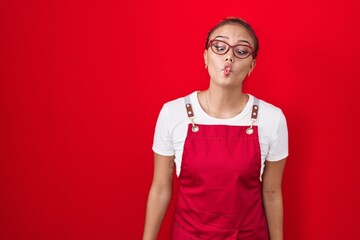 Young hispanic woman wearing waitress apron over red background making fish face with lips, crazy and comical gesture. funny expression.