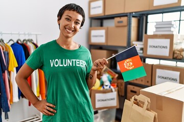 Young hispanic woman wearing volunteer uniform holding afghanistan flag at charity center