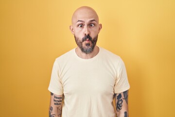 Hispanic man with tattoos standing over yellow background making fish face with lips, crazy and comical gesture. funny expression.