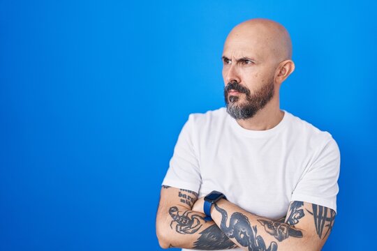 Hispanic man with tattoos standing over blue background looking to the side with arms crossed convinced and confident