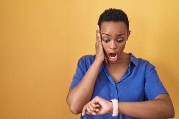 African american woman standing over yellow background looking at the watch time worried, afraid of getting late