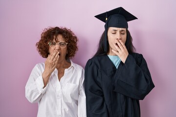 Hispanic mother and daughter wearing graduation cap and ceremony robe bored yawning tired covering mouth with hand. restless and sleepiness.