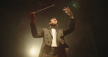 Asian symphony orchestra conductor wearing suit is directing musicians with movement of baton, isolated on black smokey background