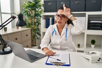Young hispanic woman wearing doctor uniform and stethoscope making fun of people with fingers on forehead doing loser gesture mocking and insulting.