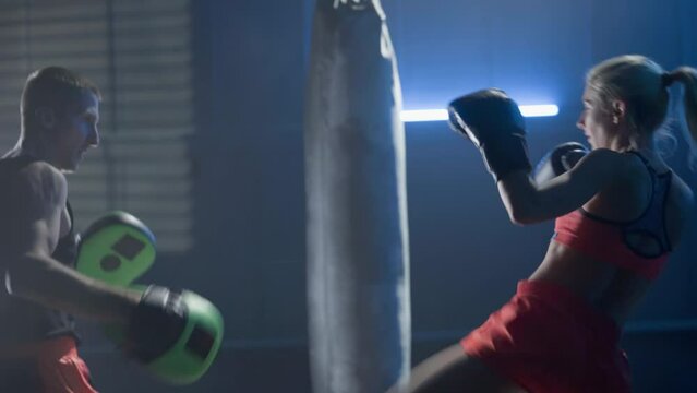 Kickboxing training. Woman in boxing gloves is boxing with her coach in dark gym with blue light. Training sparring of female kickboxer and male trainer. Female fighter practices punches and kicks.