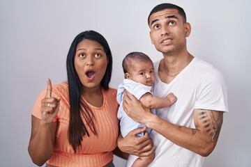 Young hispanic couple with baby standing together over isolated background amazed and surprised looking up and pointing with fingers and raised arms.