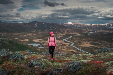Obraz premium Woman hiking in mountain landscape to Knutshoe summit in Jotunheimen National Park in Norway, view to snow-covered mountains and river and lakes