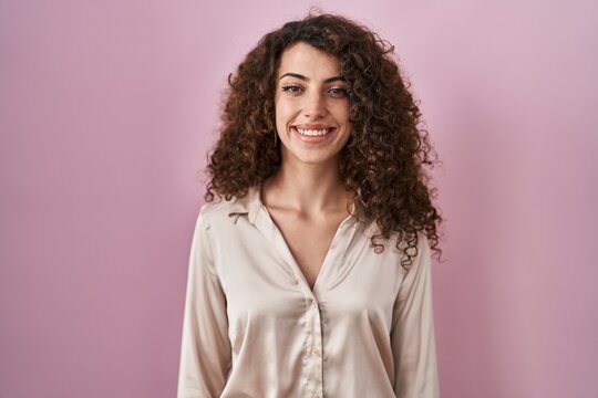Hispanic woman with curly hair standing over pink background with a happy and cool smile on face. lucky person.