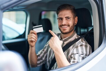Hispanic man with beard driving car holding credit card smiling happy pointing with hand and finger to the side
