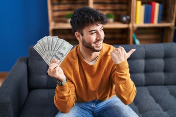 Hispanic man with beard holding 100 dollars banknotes pointing thumb up to the side smiling happy...