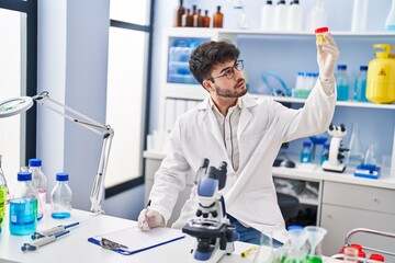 Young hispanic man scientist writing on document holding urine test tube at laboratory