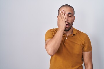 Hispanic man with beard standing over white background yawning tired covering half face, eye and mouth with hand. face hurts in pain.