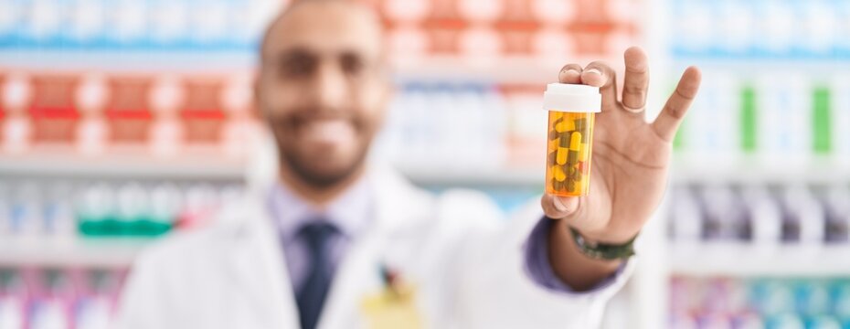 Young latin man pharmacist smiling confident holding pills bottle at pharmacy